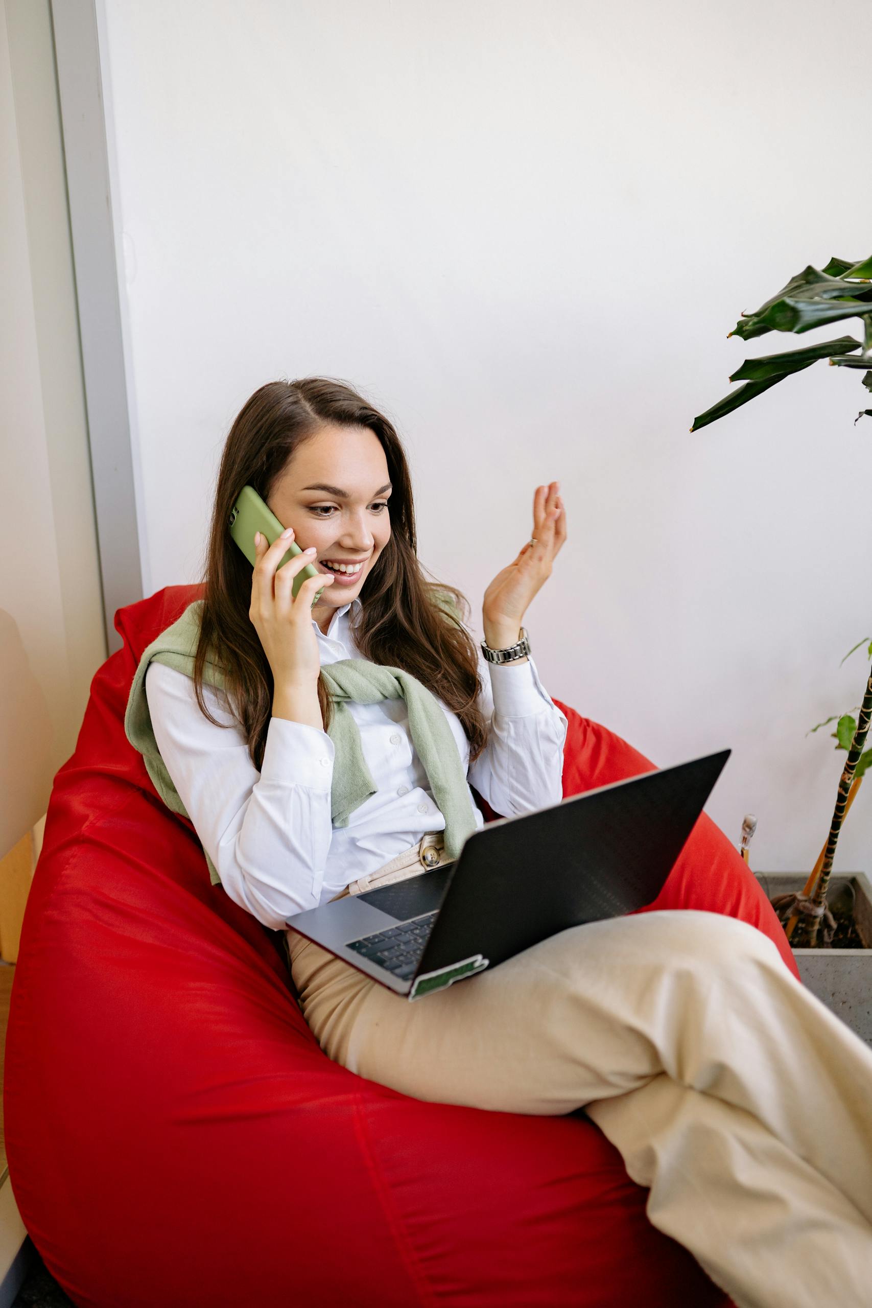 Confident young woman multitasking with a phone and laptop in a modern office setting.